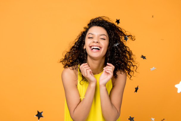 curly african american woman smiling near shiny confetti stars on orange  - 写真・画像