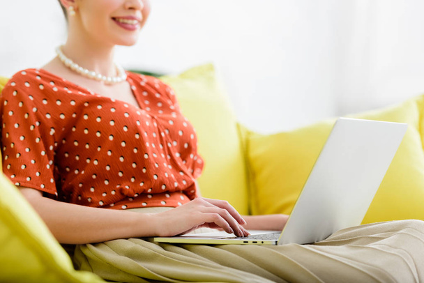 vista cortada de sorrir jovem mulher no colar de pérolas sentado no sofá amarelo e usando laptop
 - Foto, Imagem