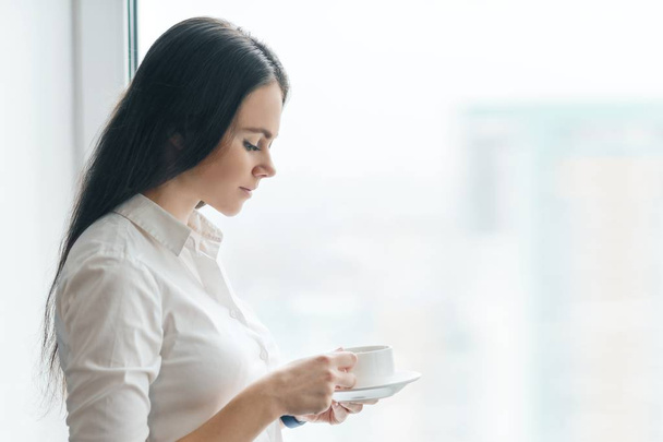 Portrait of young businesswoman in white shirt with cup of coffee, smiling woman enjoying her morning aromatic coffee, office in skyscraper cloudy day, copy space - Foto, Bild