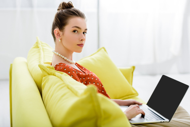 magabiztos elegáns fiatal nő, a gyöngy nyaklánc, sárga kanapén ül, és használ laptop-val üres képernyő  - Fotó, kép