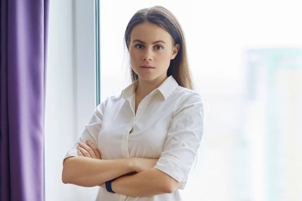 Portrait of young confident businesswoman with arms crossed near office window, female looking into camera smiling - Foto, Imagen