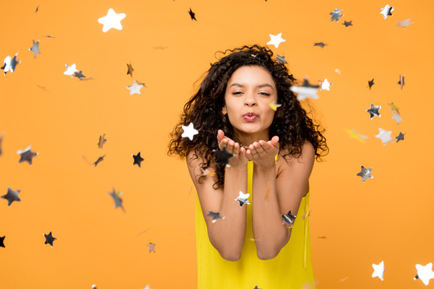 happy curly african american girl in yellow dress sending kiss near shiny confetti stars on orange  - Photo, Image