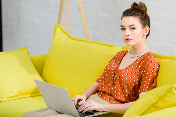 elegant young woman sitting on yellow sofa, using laptop in living room and looking at camera - Foto, Imagem
