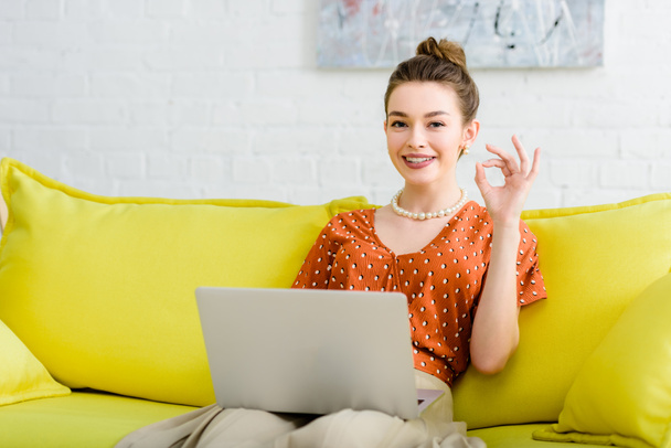 boldog elegáns fiatal nő ül sárga kanapé, használ laptop-ban nappali és rendben jel mutatja - Fotó, kép