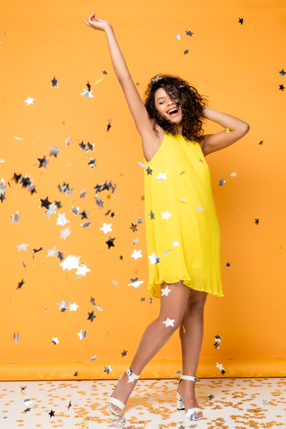 attractive curly african american girl standing near shiny confetti stars on orange  - Photo, Image