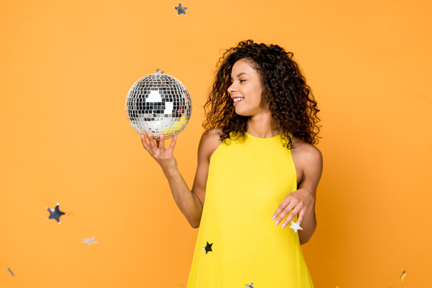 happy curly african american woman looking at disco ball near shiny confetti stars on orange  - Photo, Image