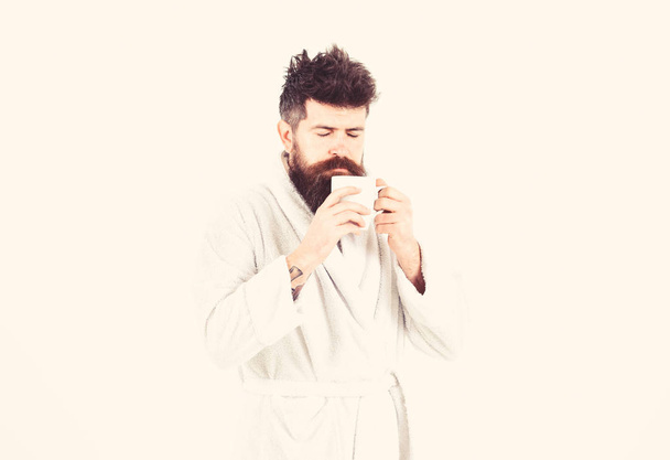 Macho drowsy, sleepy face drinks coffee in morning enjoying aroma. Morning rituals concept. Man with beard and disheveled hair stands in bathrobe, holds mug with tea or coffee, white background - Foto, Imagen