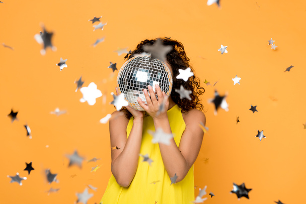 selectieve focus van Afro-Amerikaanse meisje in gele jurk voor gezicht met disco bal in de buurt van Shiny confetti sterren op oranje  - Foto, afbeelding