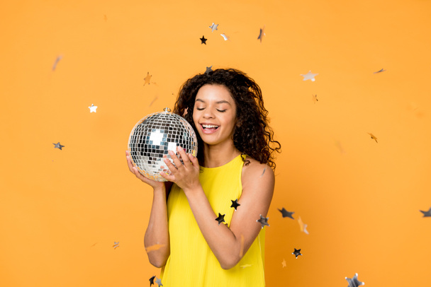 curly african american woman holding disco ball near shiny confetti stars on orange  - Photo, Image