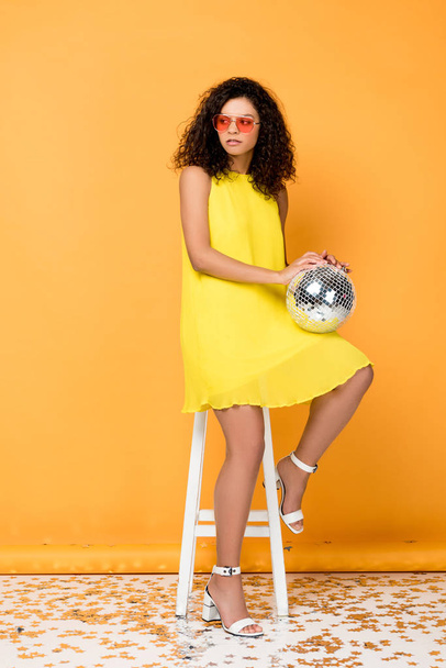 attractive curly african american woman holding disco ball while sitting on chair on orange  - Zdjęcie, obraz