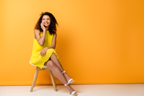 happy curly african american woman sitting in yellow dress on chair and talking on smartphone on orange - Фото, зображення