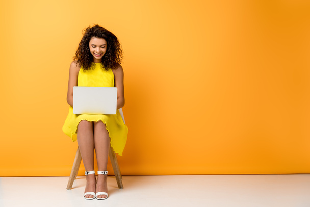 glücklich lockige Afroamerikanerin sitzt in gelbem Kleid auf Stuhl und benutzt Laptop auf orange - Foto, Bild