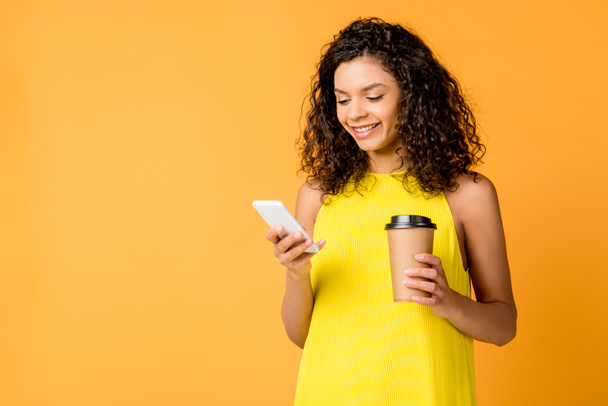 happy curly african american woman using smartphone while holding paper cup isolated on orange - Fotoğraf, Görsel