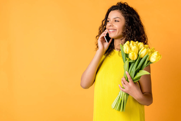cheerful curly african american woman talking on smartphone while holding yellow tulips isolated on orange - Photo, Image