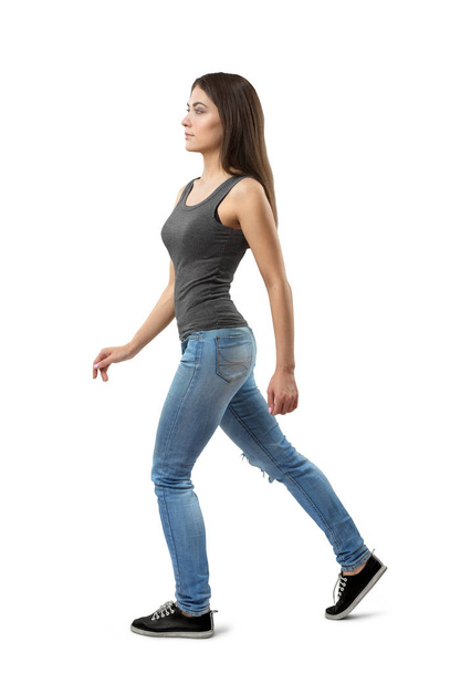 Side view of young woman in gray sleeveless top and blue jeans, with long dark hazelnut hair, walking forward on white background. - Photo, image