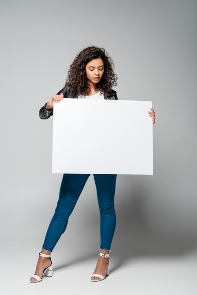 pretty curly african american girl holding blank placard while standing on grey - Foto, imagen