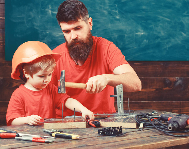 Father with beard teaching little son to use tools, hammering, chalkboard on background. Fatherhood concept. Boy, child busy in protective helmet learning to use hammer with dad - Photo, image