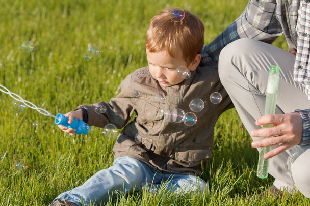 das Kind mit seinem Vater sitzt auf dem Gras und macht eine schöne Seifenblase - Foto, Bild