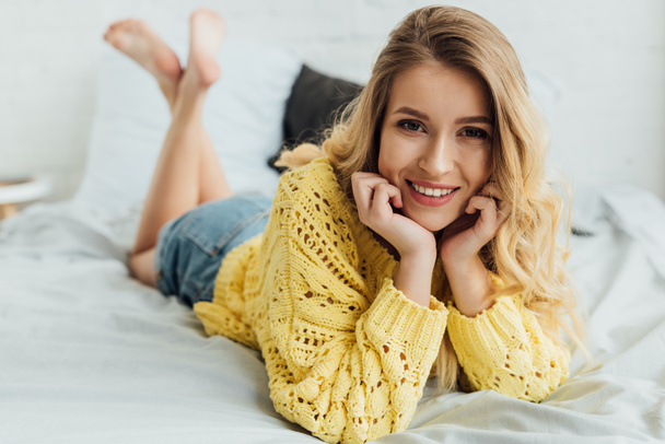 όμορφη χαμογελαστή νεαρή γυναίκα σε πλεκτό πουλόβερ που βρίσκεται στο κρεβάτι, κοιτάζοντας την κάμερα και το πρόσωπο με το χέρι με τα χέρια - Φωτογραφία, εικόνα
