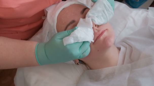 The beautician cleans the face of a beautiful girl from the remnants of the mask with the help of wet wipes. Cosmetic procedure for the rejuvenation of female skin in the beauty salon. Close-up. - Footage, Video