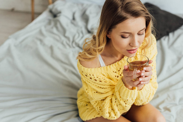 mooie jonge vrouw met ogen gesloten zittend op bed en holding kopje thee  - Foto, afbeelding
