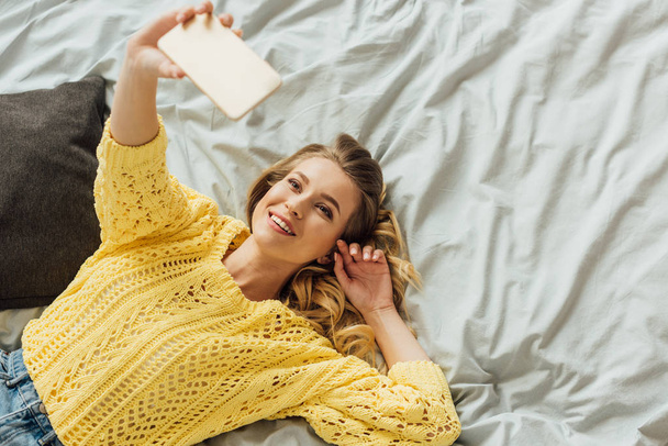 yatakta yatan güzel kız tepegöz görünümü ve smartphone üzerinde selfie alarak - Fotoğraf, Görsel