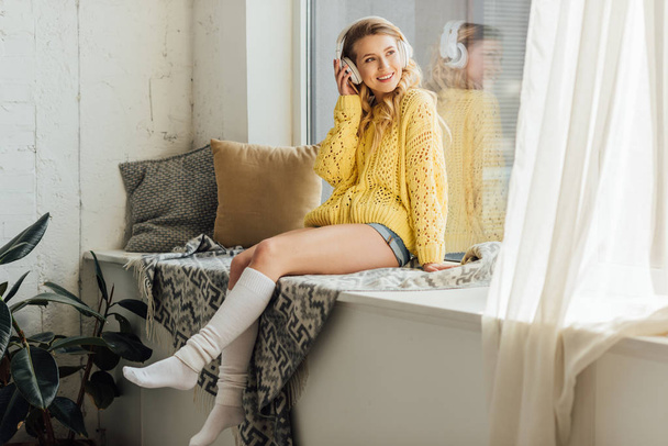 güzel gülümseyen genç kadın kulaklık pencere eşik oturan ve evde müzik dinleme - Fotoğraf, Görsel