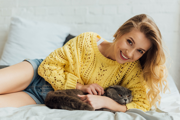 красивая улыбающаяся девушка в трикотажном свитере смотрит в камеру и обнимает шотландского кота в постели
  - Фото, изображение