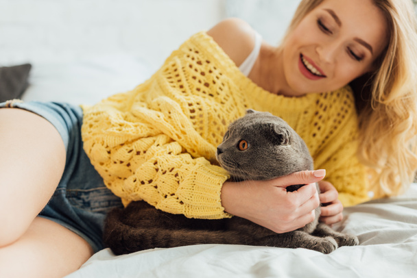 selective focus of beautiful smiling girl in knitted sweater lying in bed and hugging scottish fold cat  - Fotoğraf, Görsel