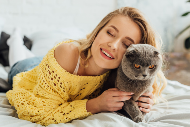 beautiful smiling girl with eyes closed lying in bed and hugging scottish fold cat  - Photo, Image