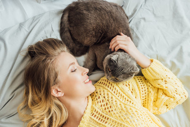 bovenaanzicht van mooie glimlachende meisje in gebreide trui strelen Schotse fold Cat terwijl liggend in bed thuis - Foto, afbeelding