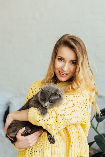 beautiful smiling girl in knitted sweater looking at camera and hugging cute scottish fold cat  - Foto, imagen