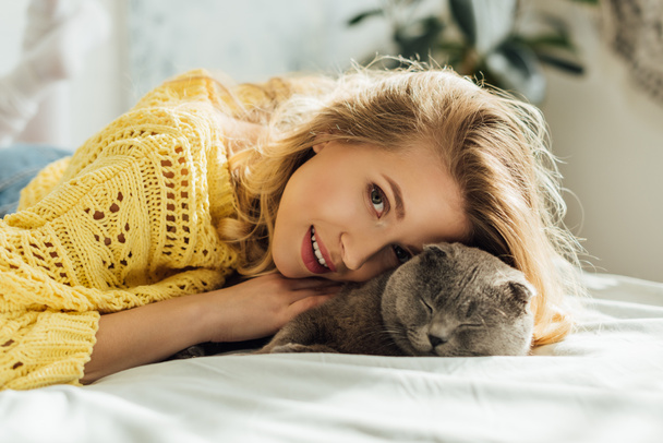 beautiful girl in knitted sweater looking at camera while lying in bed and hugging scottish fold cat  - Zdjęcie, obraz