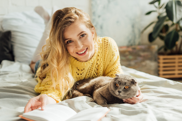 beautiful young woman looking at camera and holding book while lying in bed with scottish fold cat  - Fotó, kép