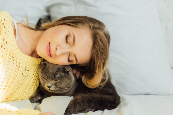 beautiful young woman with eyes closed lying in bed with adorable scottish fold cat - 写真・画像
