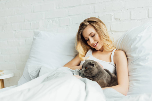 beautiful smiling girl lying in bed and hugging scottish fold cat with copy space - Photo, Image