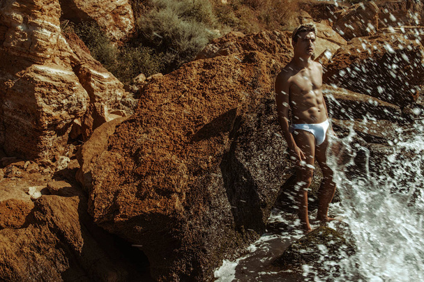 Strong face male model in white underwear on the nature background with sand, rocks and water on the beach on the sunset - Fotó, kép