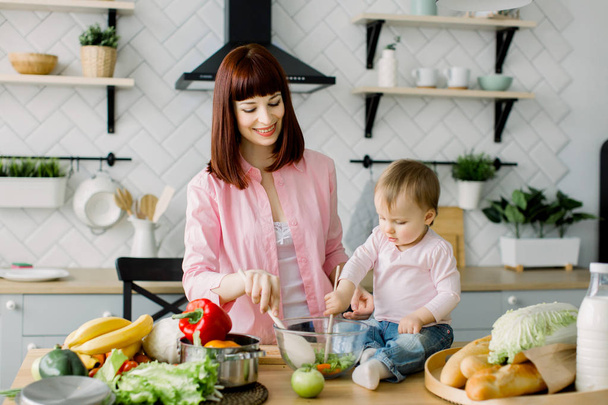 Una joven y hermosa madre con una camisa rosa está preparando una ensalada de verduras frescas en casa en la cocina, junto con su pequeña y linda hija sentada en la mesa. madre e hija
 - Foto, imagen