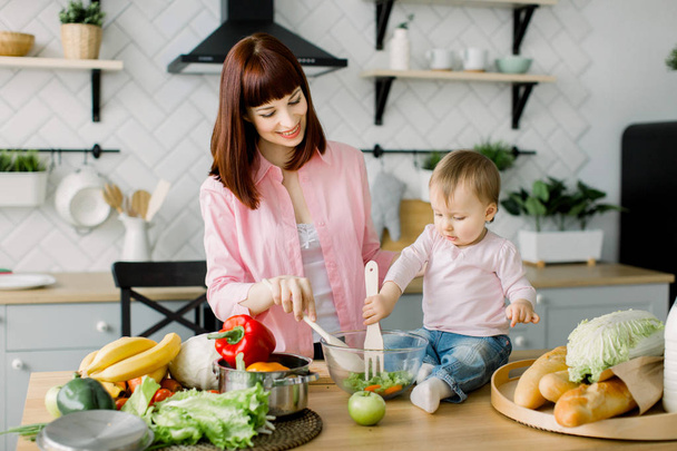 Cute little toddler baby girl holding wooden fork helping her pretty mother to mix salad at the table in the kitchen. Young beautiful woman with daughter cooking fresh salad at home. - Фото, изображение