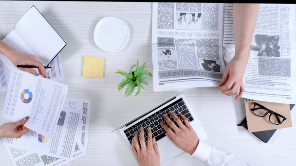 bovenaanzicht van ondernemers met behulp van laptop, holding van papers en schrijven in notitieblok, krant lezen en koffie drinken - Video