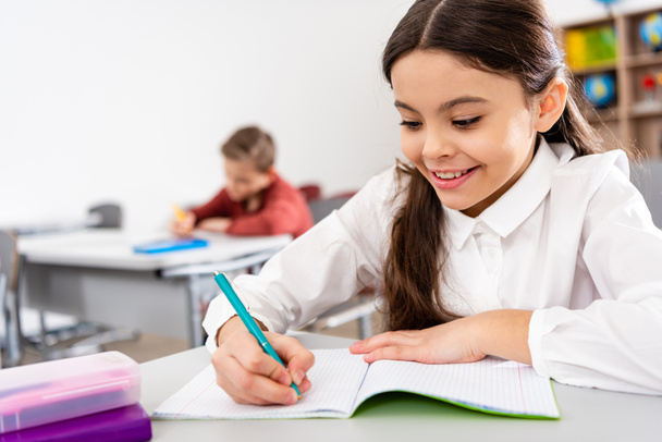 Smiling schoolgirl writing in notebook during lesson in classroom - Photo, Image