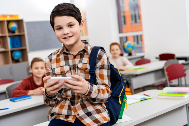 Smiling pupil in checkered shirt with backpack using smartphone in classroom - Photo, Image