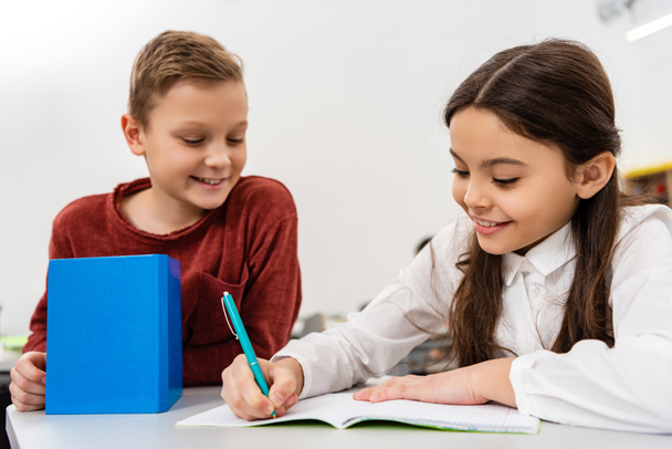 Glimlachend school meisje praten met vriend tijdens het schrijven in notebook in de klas - Foto, afbeelding