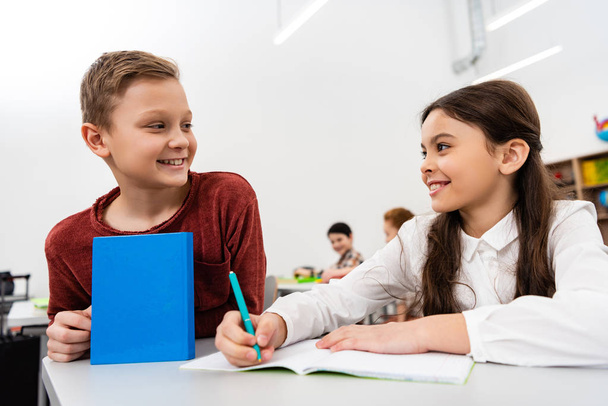 Усміхнена школярка розмовляє з другом під час написання в блокноті в класі
 - Фото, зображення
