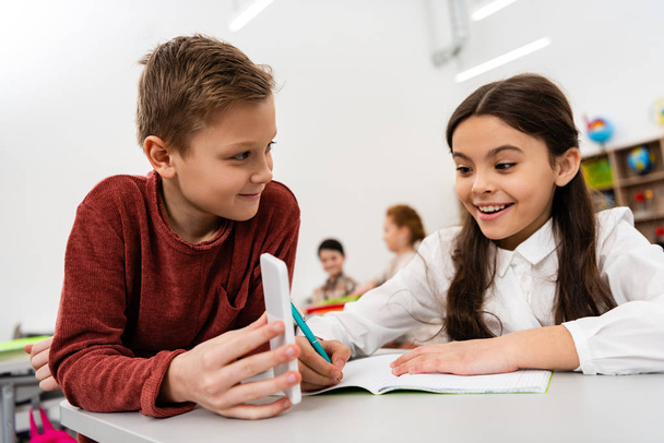 Smiling schoolboy showing smartphone to friend at desk in classroom - Foto, Imagen
