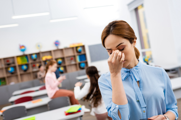 Tired teacher in blue blouse standing in front of desks and touching nose - Фото, зображення