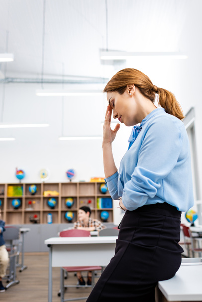 Väsynyt opettaja sinisessä puserossa seisomassa pöydän lähellä luokkahuoneessa
 - Valokuva, kuva