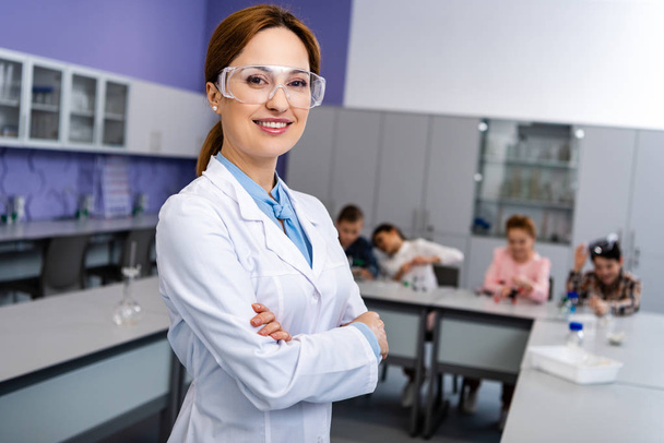 Lachende leraar in beschermende bril staande met gekruiste armen in de voorkant van de leerlingen tijdens de chemie les - Foto, afbeelding