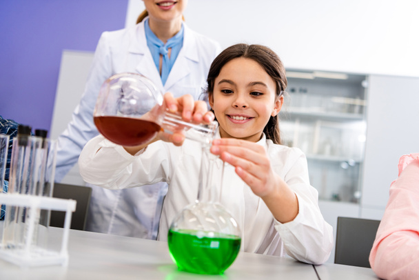 Colegiala sonriente con vasos haciendo experimento químico durante la lección de química
 - Foto, Imagen