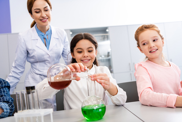 Colegiala sonriente con vasos haciendo experimento químico durante la lección de química
 - Foto, Imagen